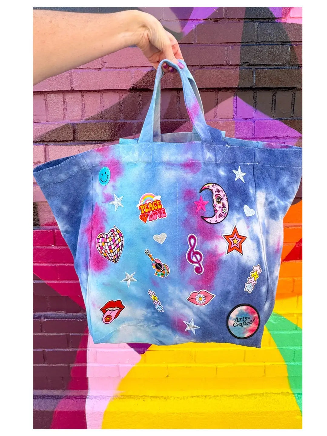Tie-dye tote bag with colorful iron-on patches held against a vibrant mural.